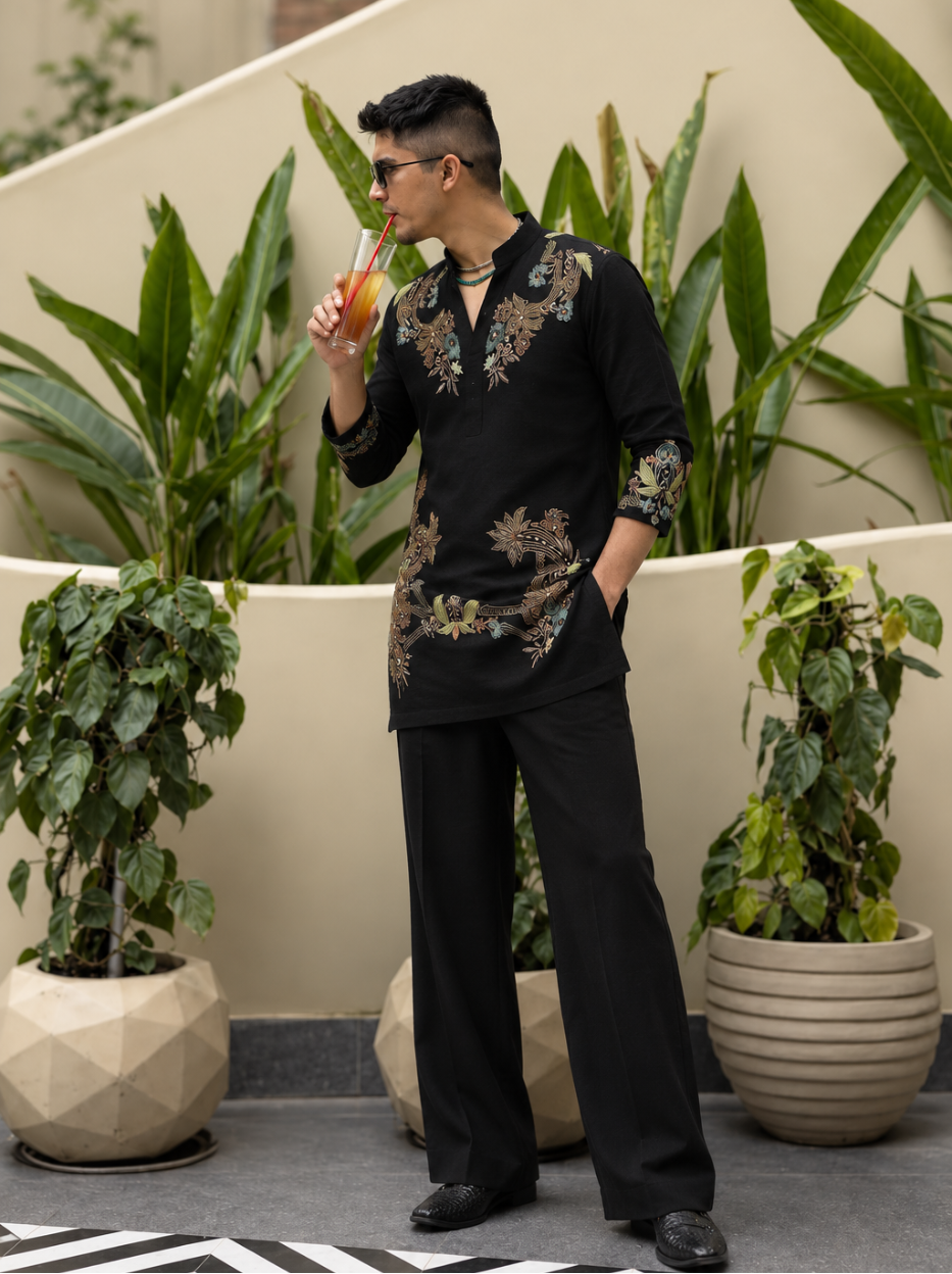 A man wearing a black embroidered traditional Chinese-style long-sleeve shirt and matching trousers, standing in an outdoor patio setting with potted plants and drinking from a glass with a straw.